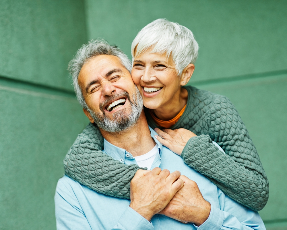 Smiling elderly couple embracing against green wall, enjoying life with restored vision after surgery